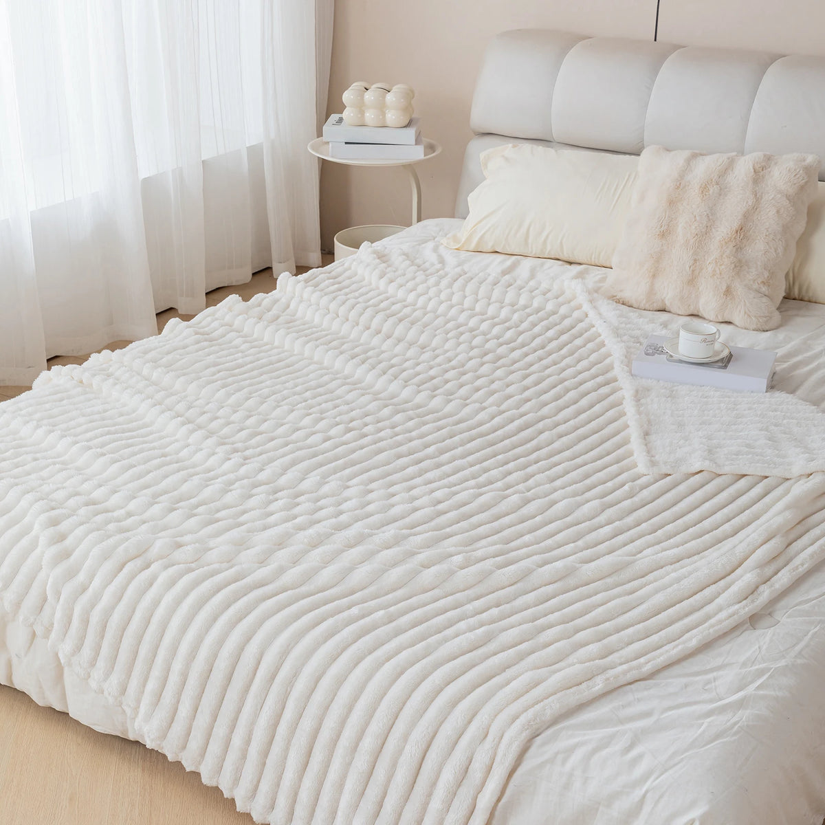 A white knitted blanket is draped over a bed, next to pillows and a shelf of rolled towels.