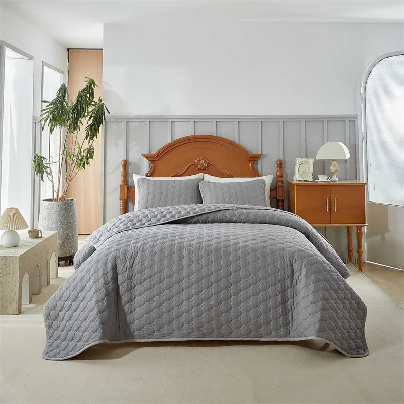 A light gray quilted bedspread set is displayed on a bed with a wooden headboard in a bright bedroom.