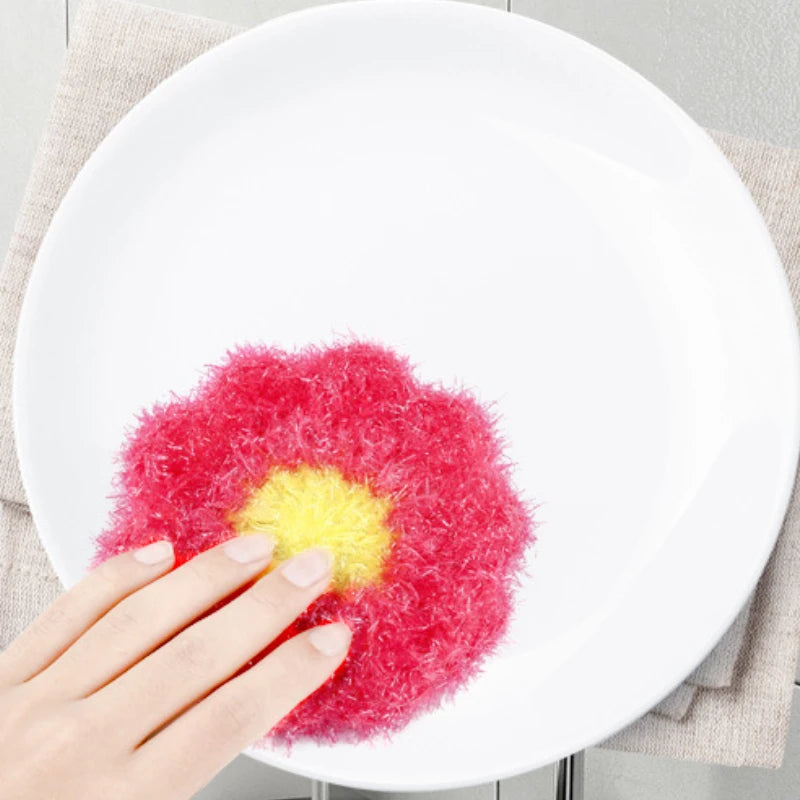 A hand places a bright pink and yellow flower-shaped kitchen dish scrubber sponge on a white plate.