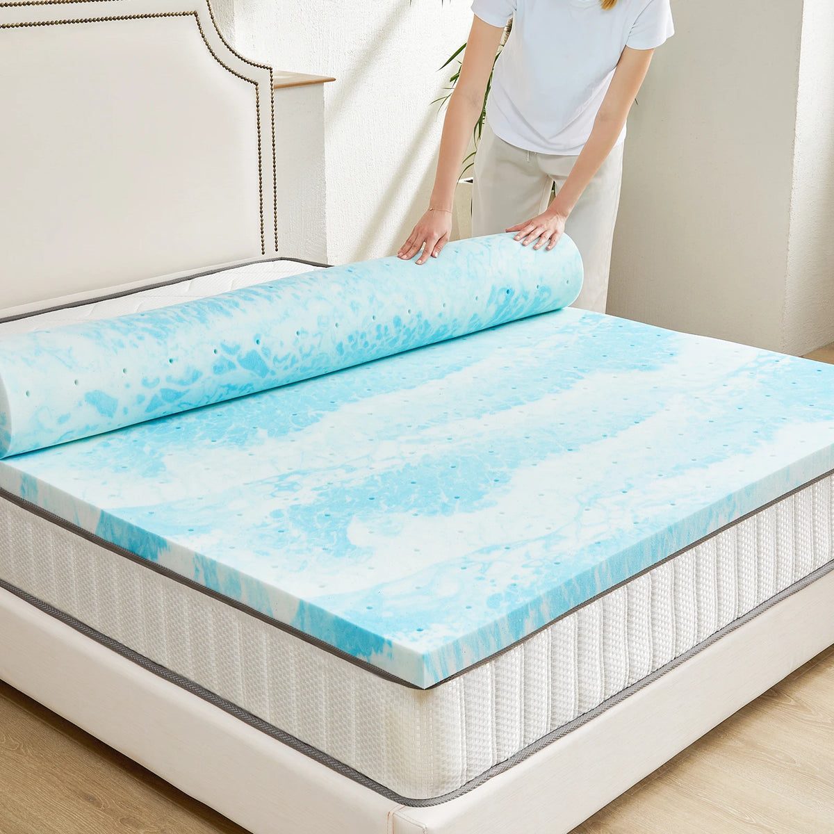 A person is placing a blue and white cooling memory foam mattress topper onto a white bed frame.