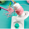 A white hand-operated wool yarn winder is shown with pink yarn being wound onto it on a teal background.