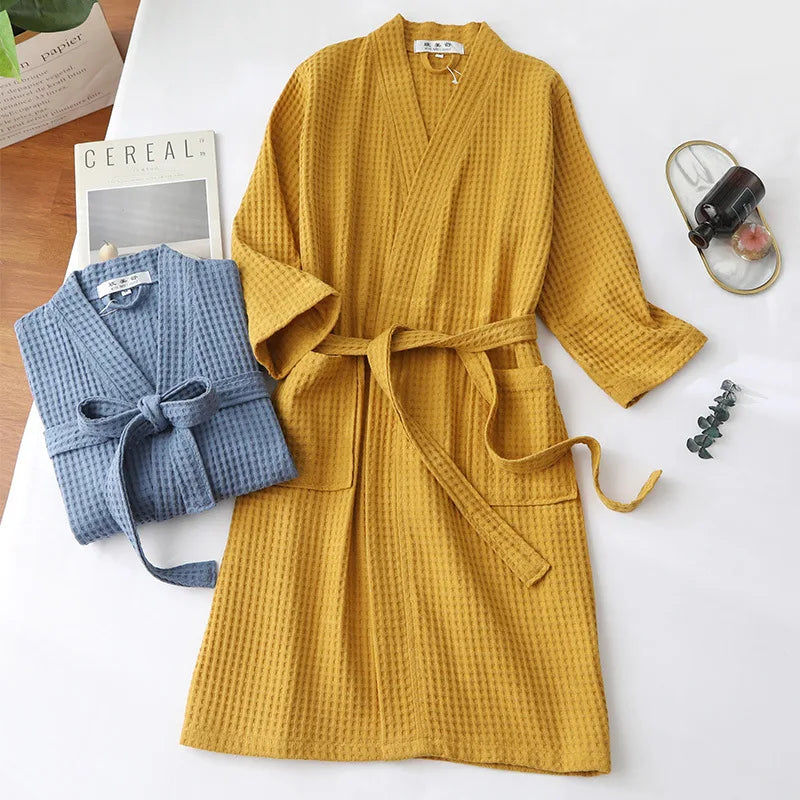 A mustard yellow cotton terry robe hanging on a white surface next to a folded blue robe and a box.