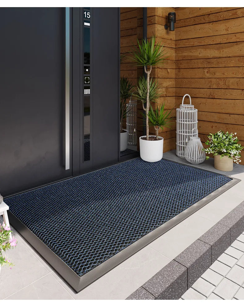 A dark blue non-slip rubber doormat placed on a concrete walkway near a wooden wall and potted plant.