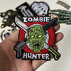 A hand holds a black embroidered patch featuring a green zombie head with crossed hunting knives and the word HUNTER.