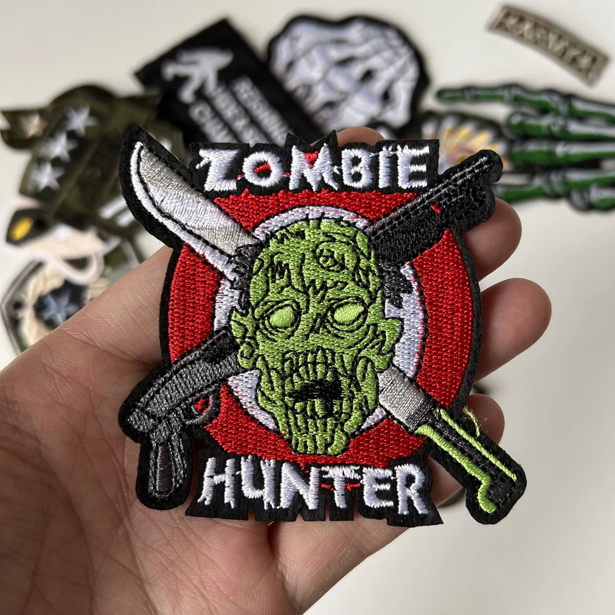 A hand holds a black embroidered patch featuring a green zombie head with crossed hunting knives and the word HUNTER.