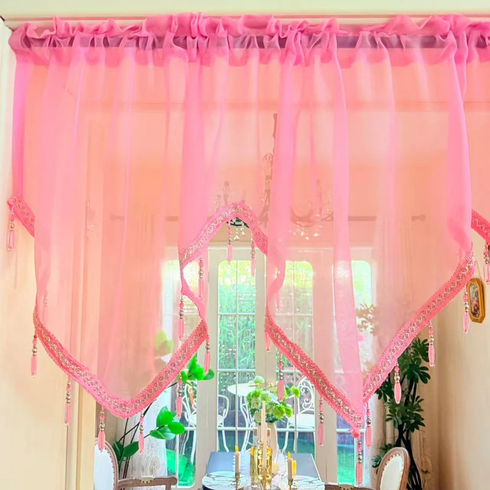 Pink sheer curtain with lace trim and tassels hanging in a window frame.