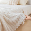 A hand is touching a fluffy white blanket draped over a bed, showing its soft texture.