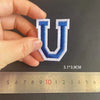 A hand holds a blue embroidered letter U patch against a ruler showing its dimensions.