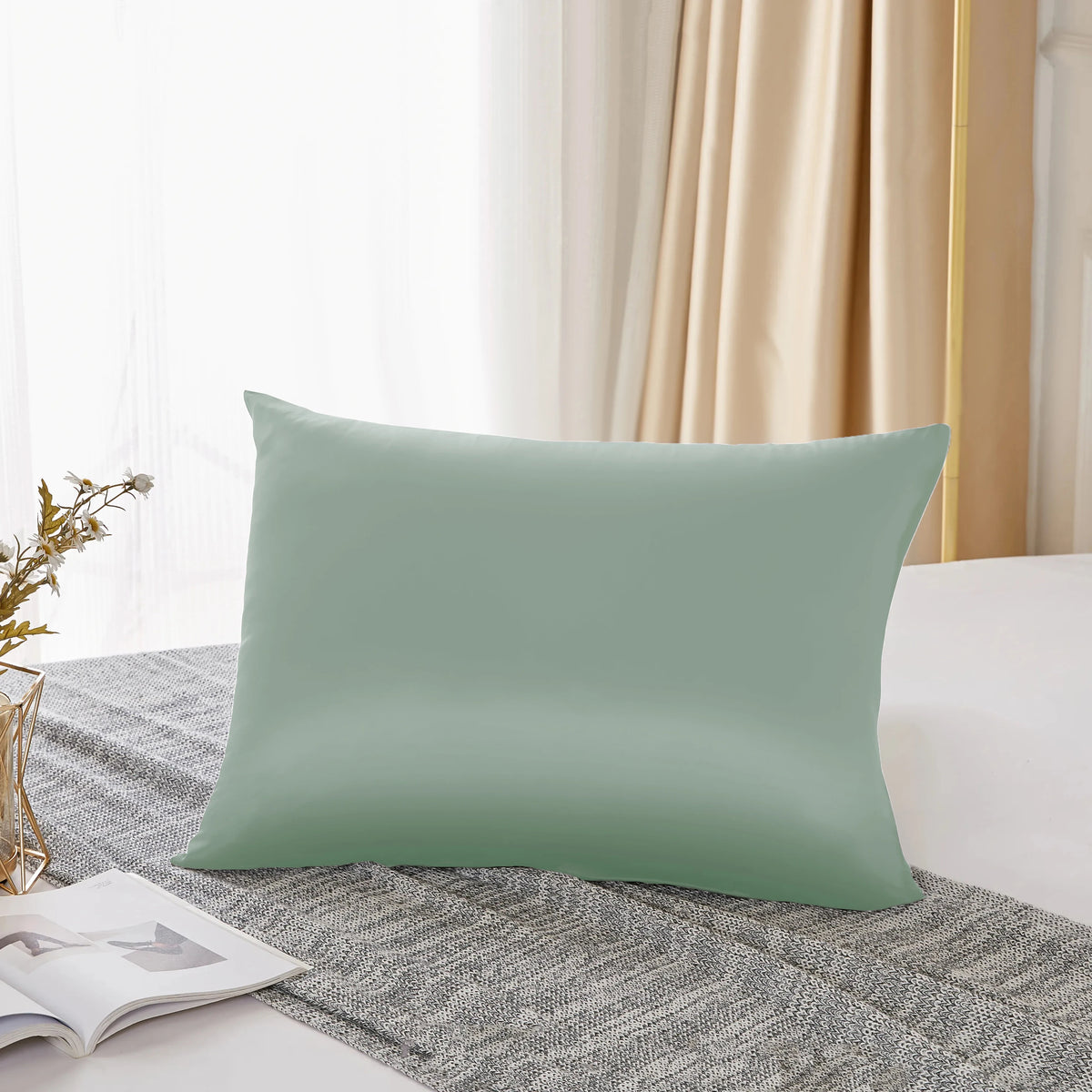 A light sage green silk pillowcase resting on a gray textured fabric near a window.
