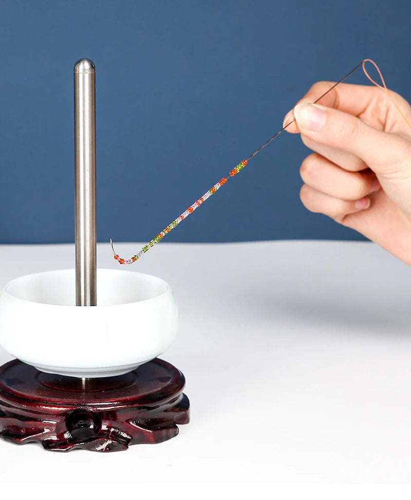 A hand is using a thin wire or needle to thread beads onto a stainless steel tool resting on a dark base.
