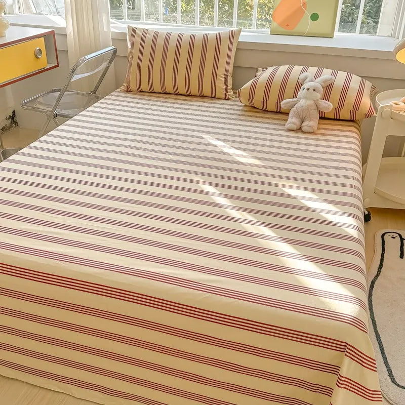 A bed dressed with a striped beige and red organic cotton flat sheet and pillows, featuring a small stuffed animal.