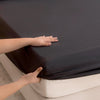 A hand is adjusting a dark gray waterproof fitted bed sheet on a white mattress.