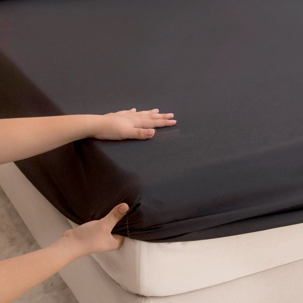 A hand is adjusting a dark gray waterproof fitted bed sheet on a white mattress.