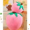 A pink plush strawberry throw pillow with green leaves is displayed on a wooden surface.
