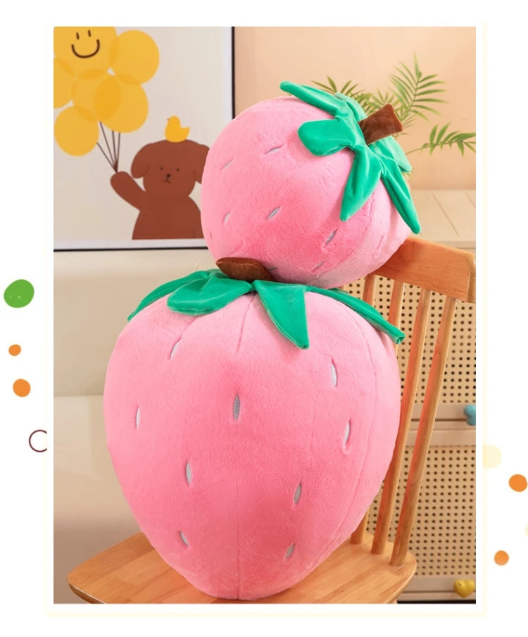 A pink plush strawberry throw pillow with green leaves is displayed on a wooden surface.