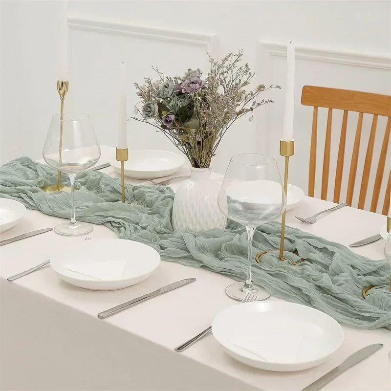 A white table setting featuring a sage green gauze table runner, white plates, silverware, and tall candles.