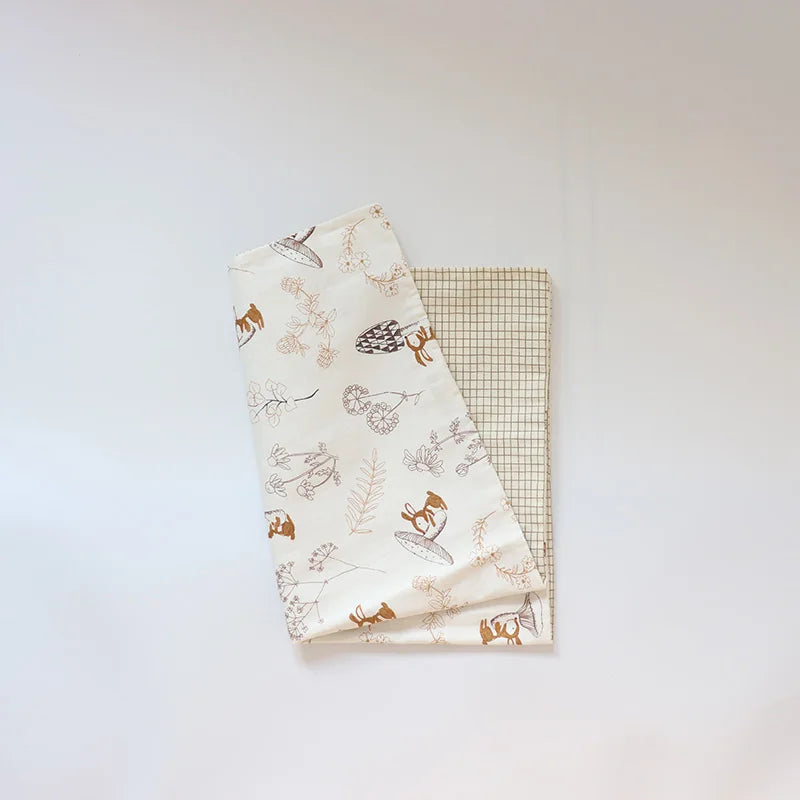 A white fabric baby pillow cover featuring small brown and green nature-themed illustrations on a light green plaid backgroun