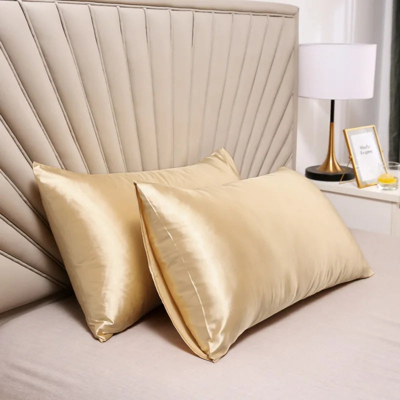 Two golden satin silk pillow covers resting on a beige bed with a padded headboard and a lamp.