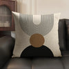 A beige cushion cover featuring black geometric lines and a circular brown accent on a dark gray sofa.