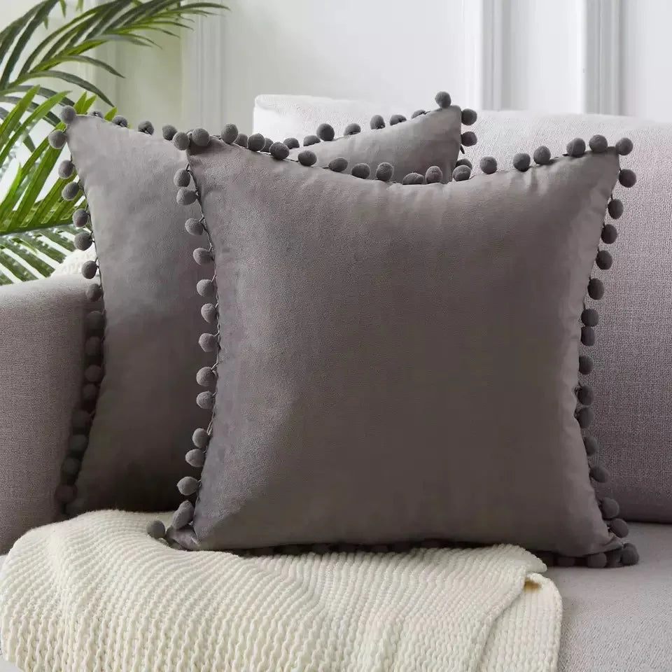 A square gray velvet cushion with decorative tasseled trim resting on a light cream knitted blanket.