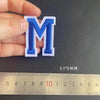 A hand holds a blue alphabet embroidery patch letter M next to a ruler showing its dimensions.