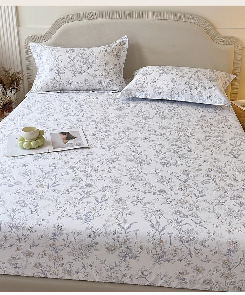 A floral printed cotton fitted sheet with a white background is displayed on a bed with a upholstered headboard.