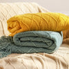 A stack of knitted tassel throw blankets in yellow and blue-green colors resting on a beige textured fabric.