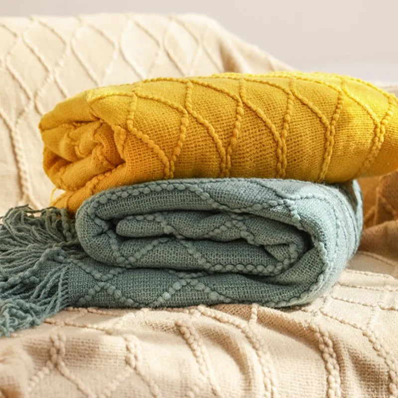 A stack of knitted tassel throw blankets in yellow and blue-green colors resting on a beige textured fabric.