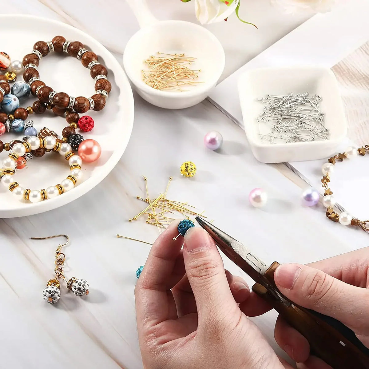 Hands are using tweezers to place stainless steel ball head pins onto jewelry components and loose beads on a white surface.