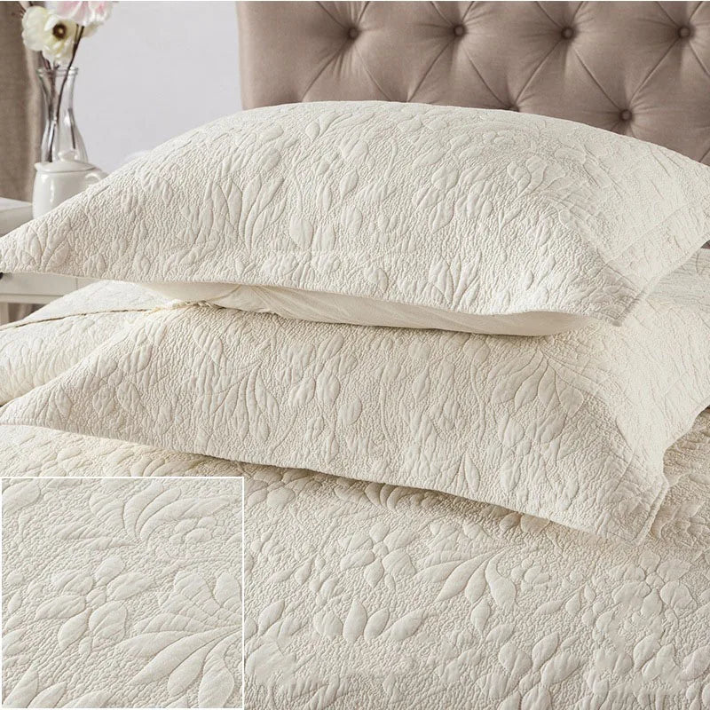 A set of white embroidered cotton quilted pillow covers stacked on a bed with a taupe upholstered headboard.