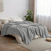A gray cotton gauze blanket is draped over a bed in a bright room next to a window.