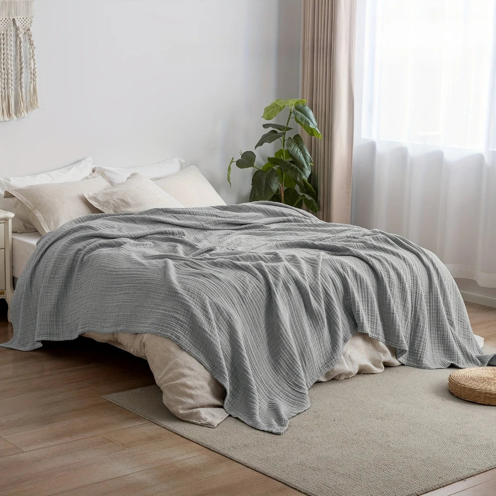 A gray cotton gauze blanket is draped over a bed in a bright room next to a window.