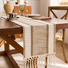A beige macrame tassel table runner is draped across a wooden table, set in a bright farmhouse interior.