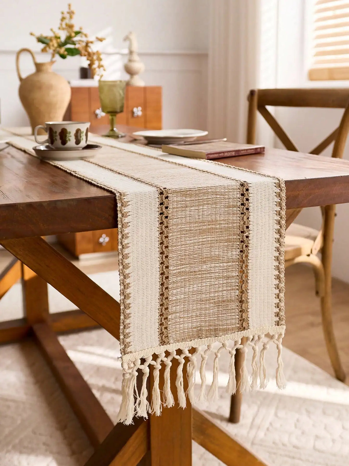 A beige macrame tassel table runner is draped across a wooden table, set in a bright farmhouse interior.