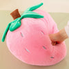 A pink plush strawberry throw pillow with green leaves and a brown stem is being held by a hand.