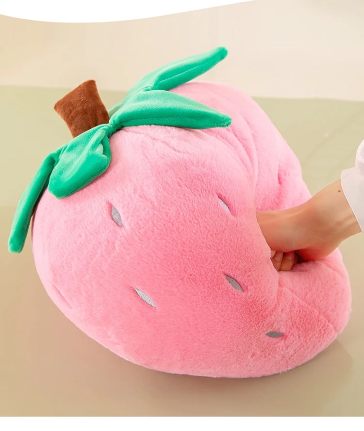 A pink plush strawberry throw pillow with green leaves and a brown stem is being held by a hand.