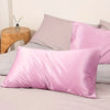 A pink satin pillowcase with an envelope closure is displayed on a bed with other pillows and a wooden side table.