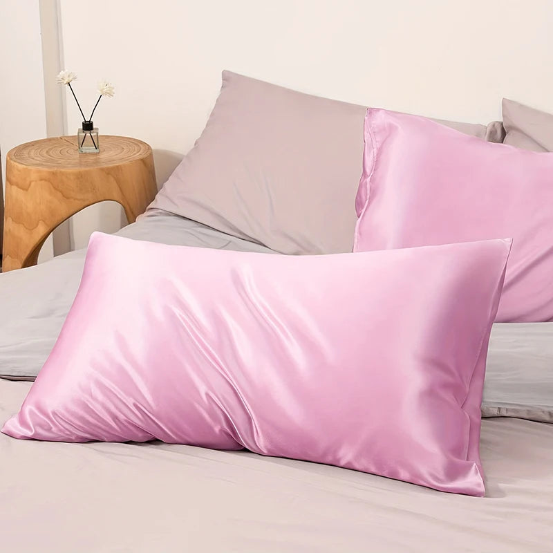 A pink satin pillowcase with an envelope closure is displayed on a bed with other pillows and a wooden side table.