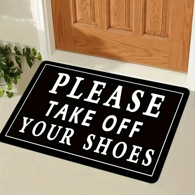 A black doormat with white text that reads Please Take Off Your Shoes placed in front of a wooden door.
