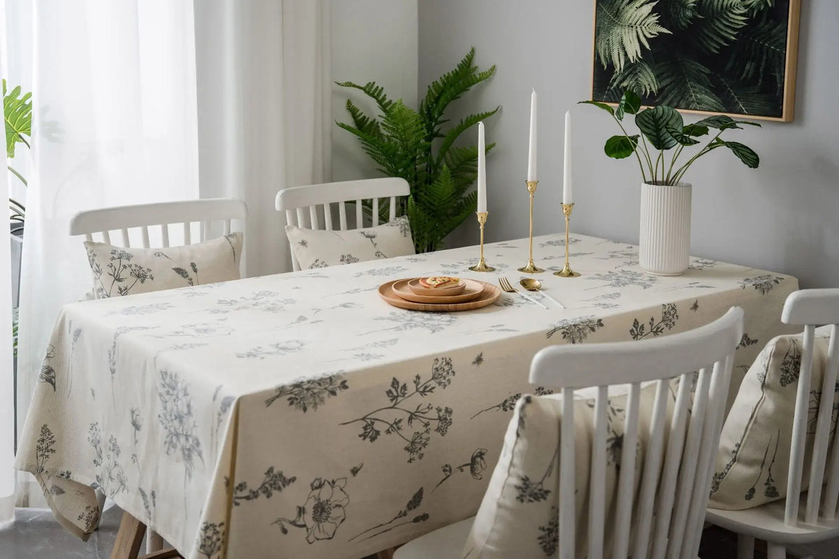 A long dining table set with a botanical print linen tablecloth, white wooden chairs, and green foliage decor.