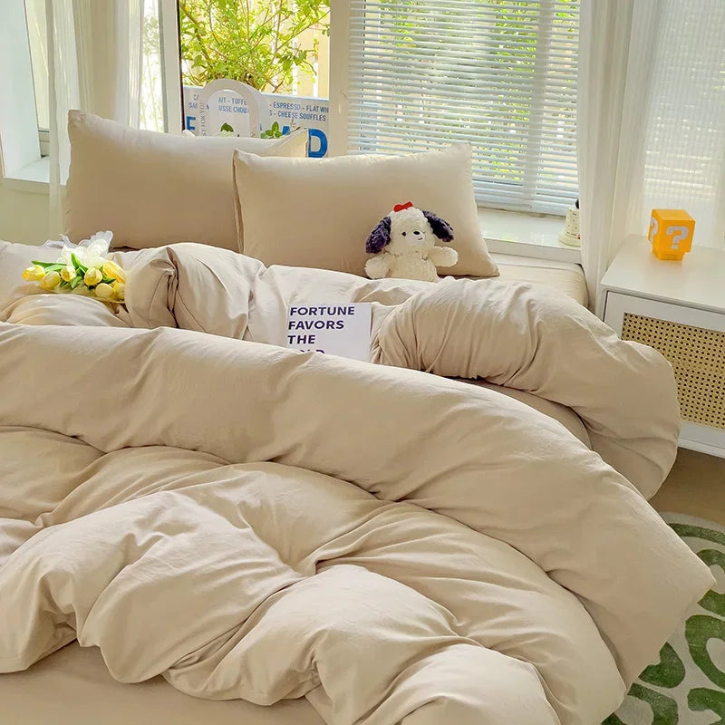 A beige solid cotton bed linen set with pillows and a duvet on a bed near a window.