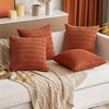 A collection of textured, rust-colored corduroy throw pillow covers and a beige knitted throw blanket on a white sofa.
