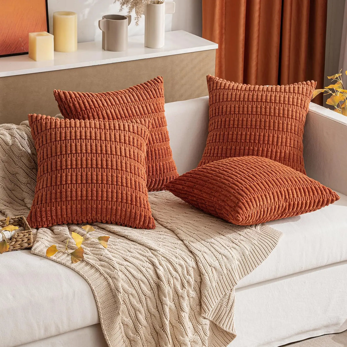 A collection of textured, rust-colored corduroy throw pillow covers and a beige knitted throw blanket on a white sofa.