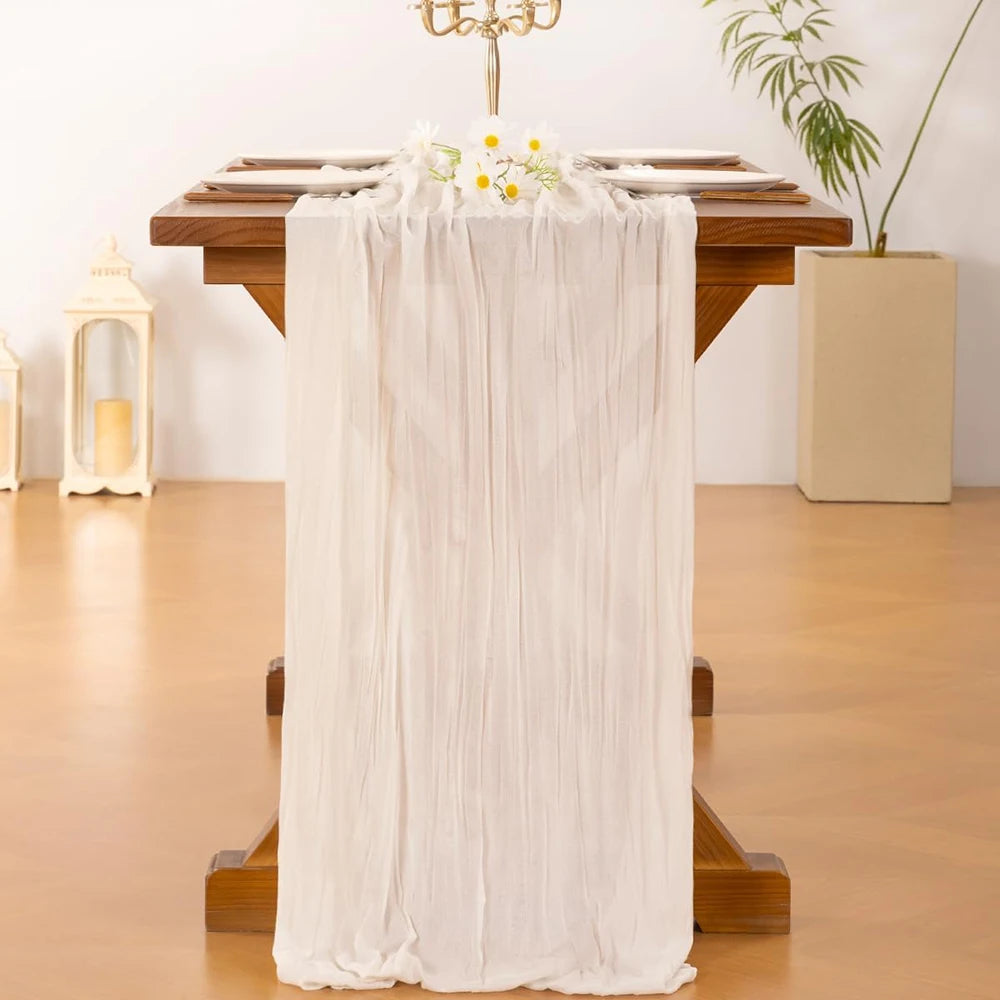A cream cheesecloth table runner with white flowers is draped over a wooden table for boho wedding decor.