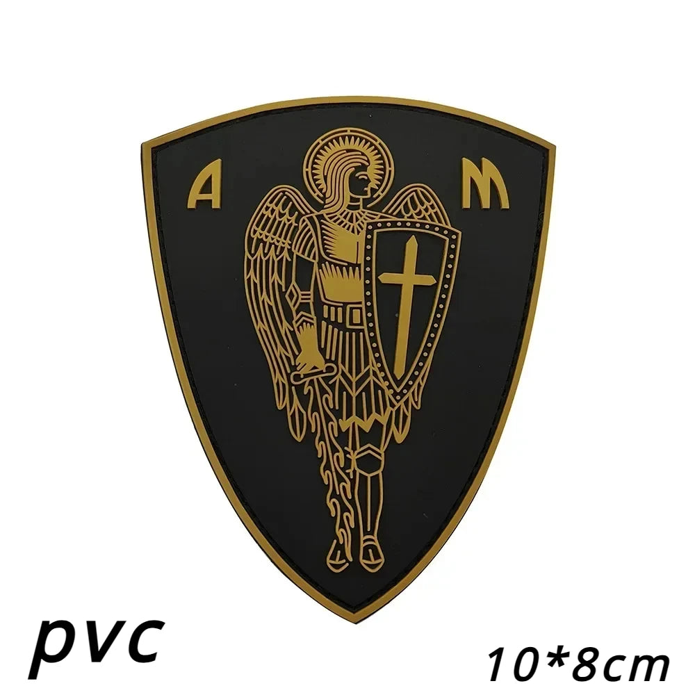A black shield patch with gold embroidery featuring an angel and a cross, labeled PVC.