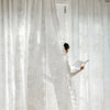 A person in a white dress is adjusting a sheer, white vintage ruffled lace embroidered curtain.