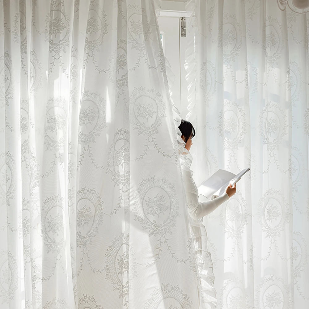 A person in a white dress is adjusting a sheer, white vintage ruffled lace embroidered curtain.