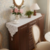 A dark wood sideboard with a lace table runner, a mirror, and floral arrangements on top.