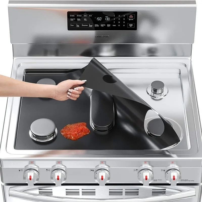 A hand places a black reusable liner protector onto the cooking surface of a stainless steel gas stove.