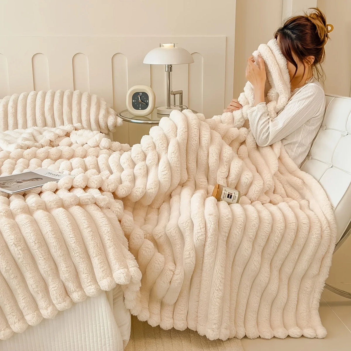 A fluffy cream fleece throw blanket draped over a white bed, next to a lamp and clock.